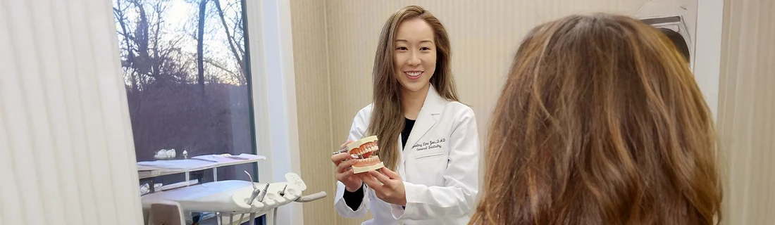 Yanling Lina Yue Showing Model Teeth to a Patient Smiling About Us