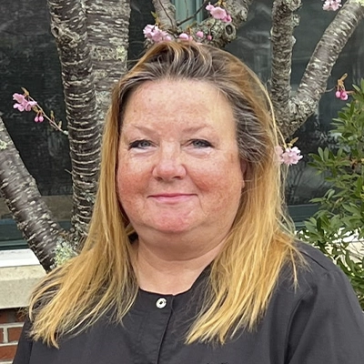 Headshot of Debbie wearing a dark top standing near trees and greenery.